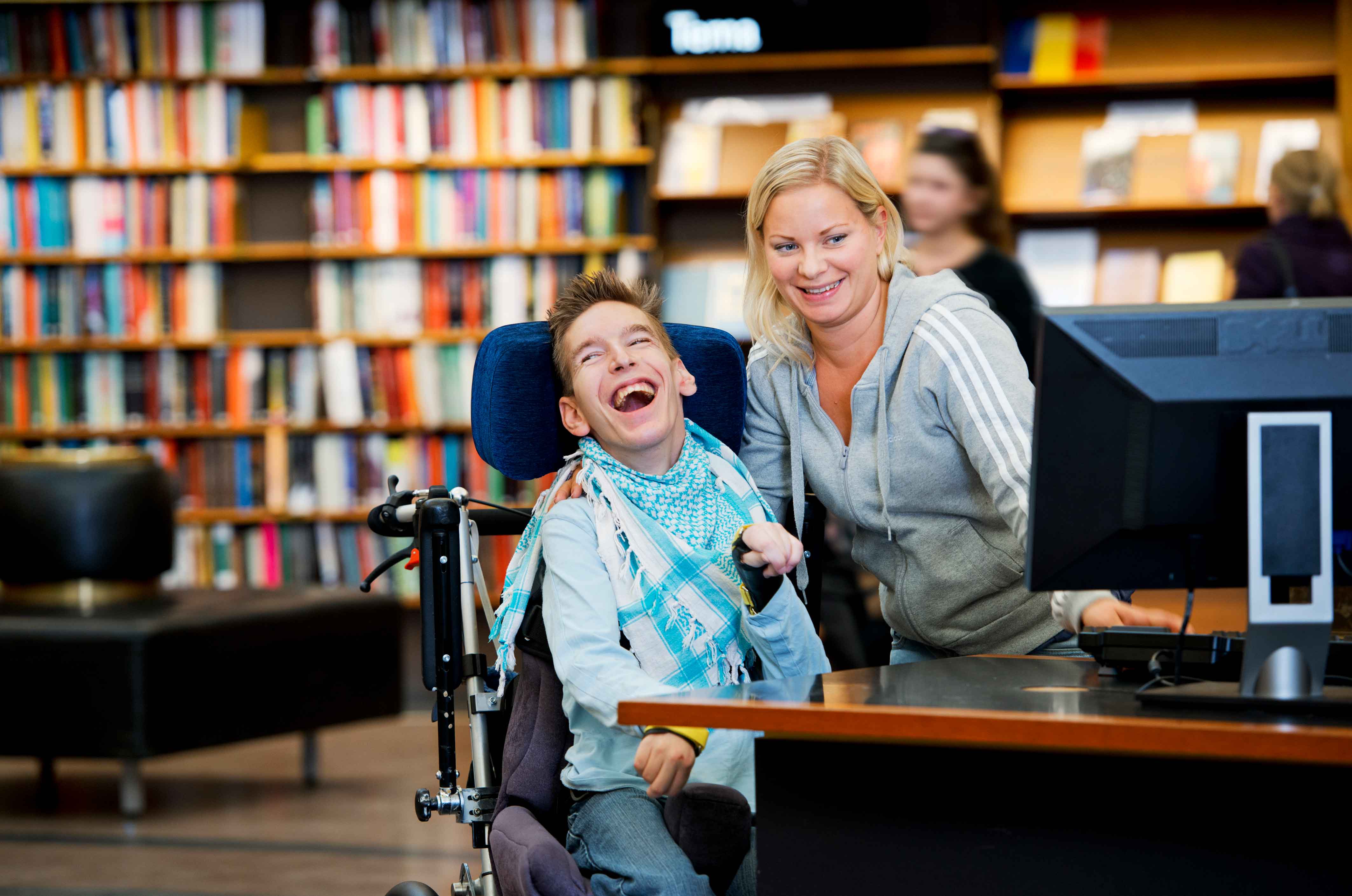 En ung man i rullstol skrattar tillsammans med en kvinna som visar honom något på en datorskärm. Omgivningen är ett bibliotek